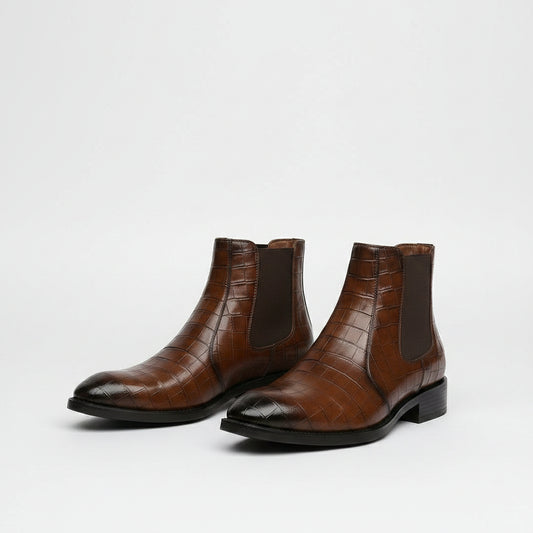 A person seated on a red chair wearing brown embossed leather Chelsea boots with dark elastic side panels and white socks, against a light wood floor and paneled wall.