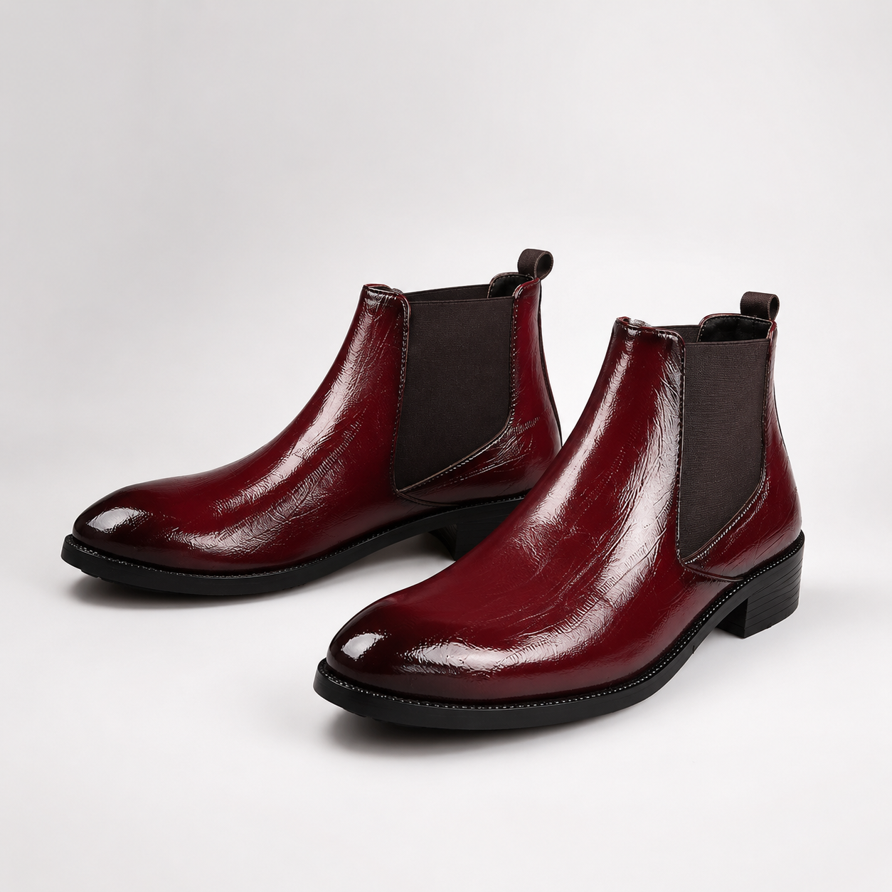 A pair of glossy deep burgundy ankle boots with dark elastic side panels, shown on a clean light gray studio background.