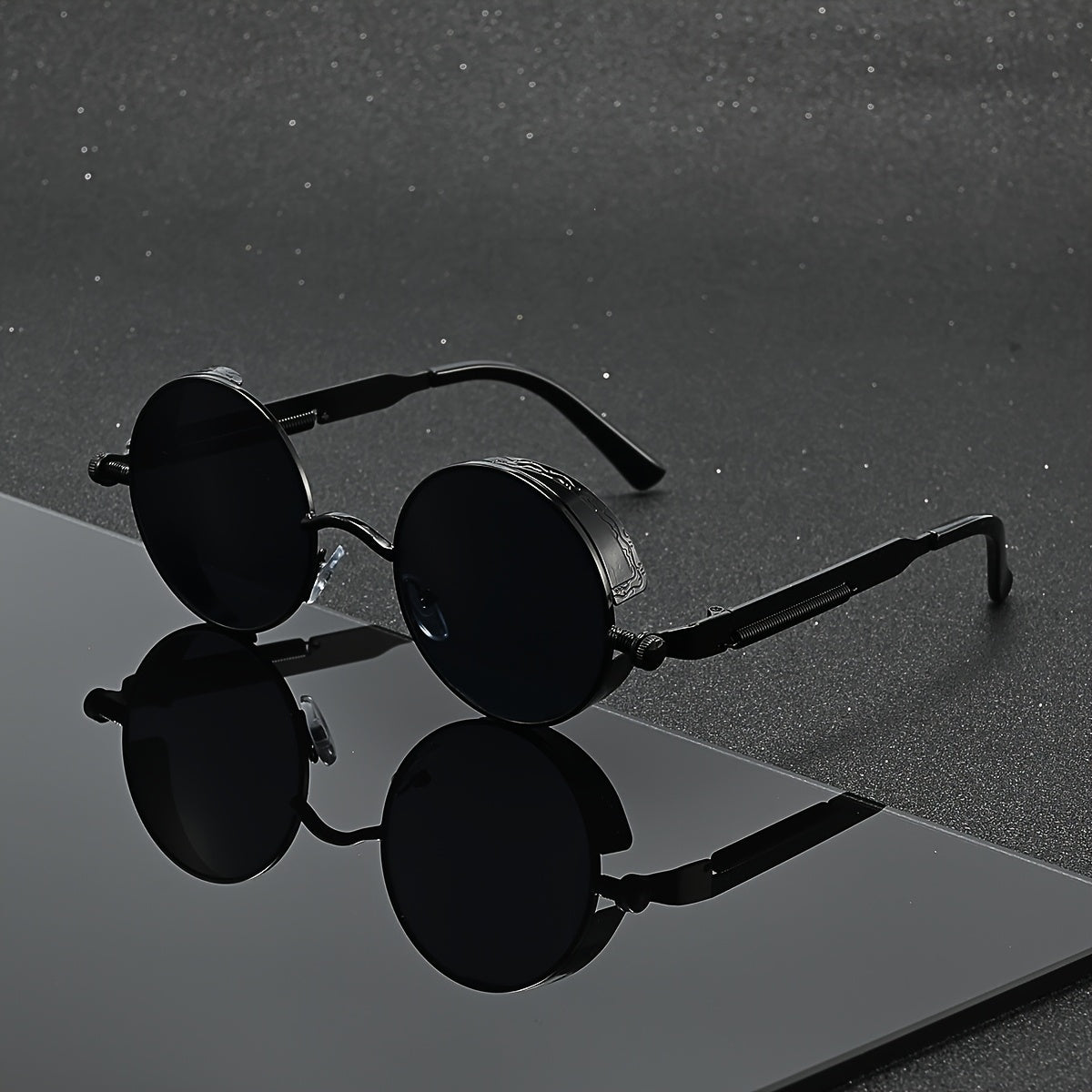 Round black sunglasses with a unique circular lens design and thin metal temples, displayed on a reflective dark surface against a textured gray background.