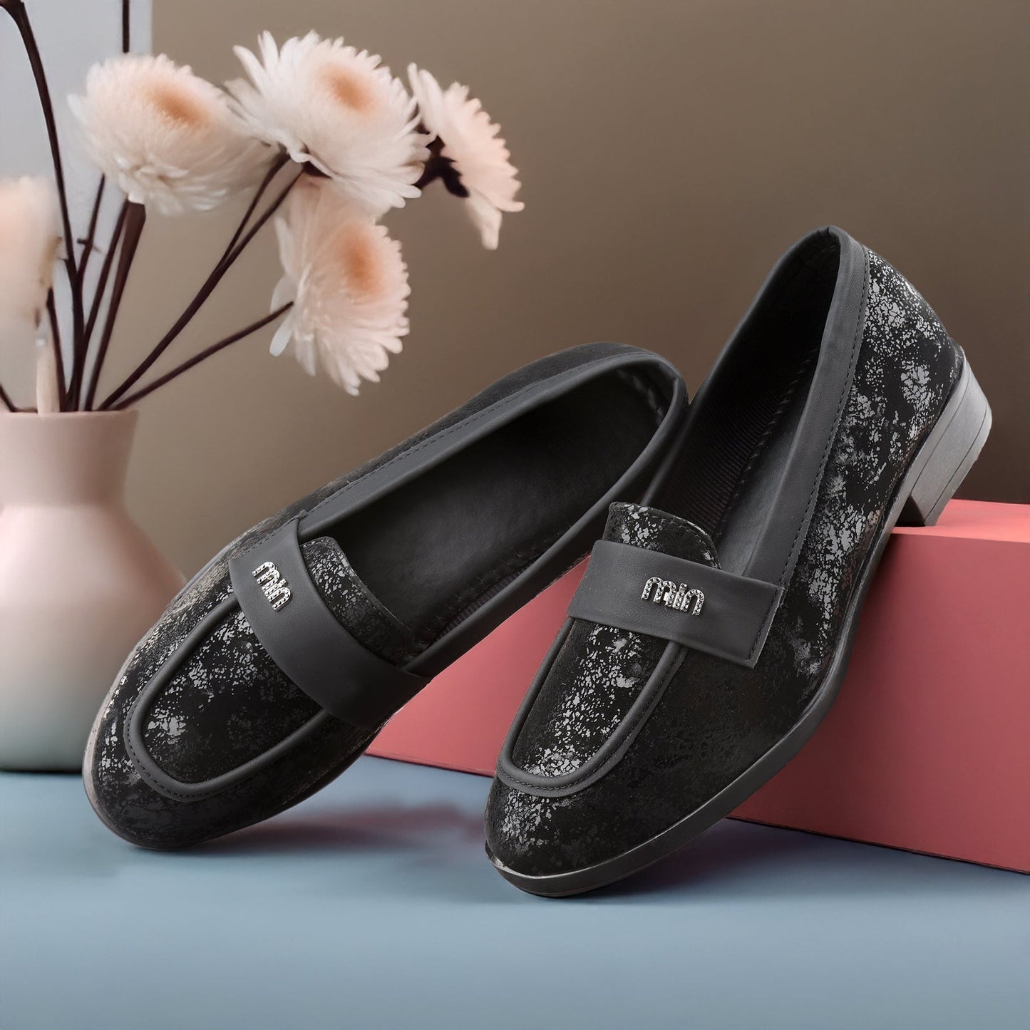 Black glittery loafers with a visible brand logo on a pink box with flowers in the background.