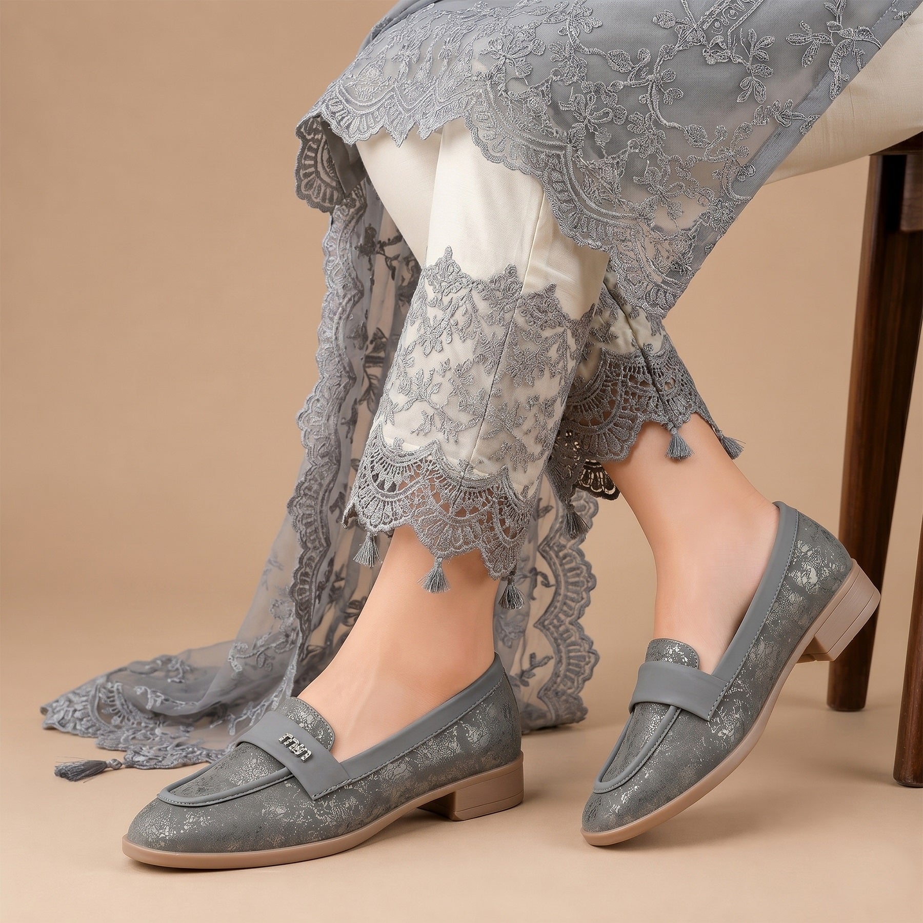 A person seated on a chair wearing gray lace-up loafers with a textured floral pattern, paired with white pants and a sheer gray lace shawl against a beige studio background.