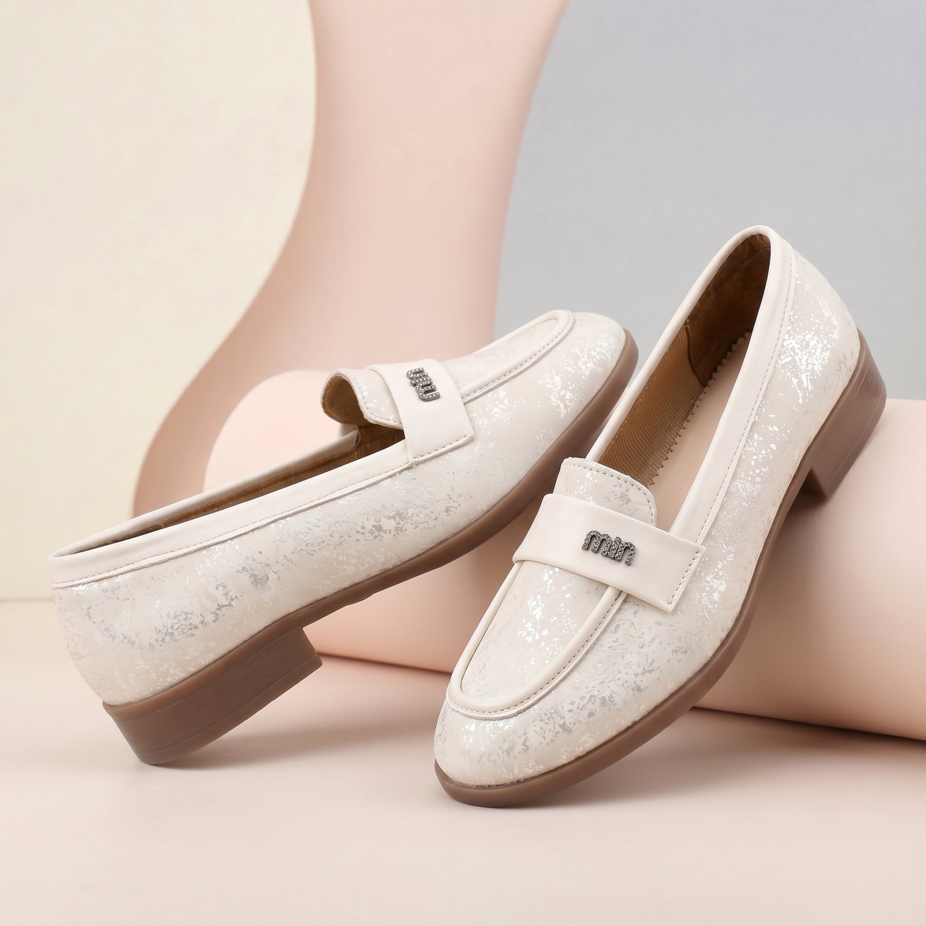A pair of women's loafers in a textured off-white fabric with a subtle glitter finish, featuring a low block heel and a decorative strap with the word 'mim' in rhinestones. They are displayed on a minimalist beige and light gray curved background.