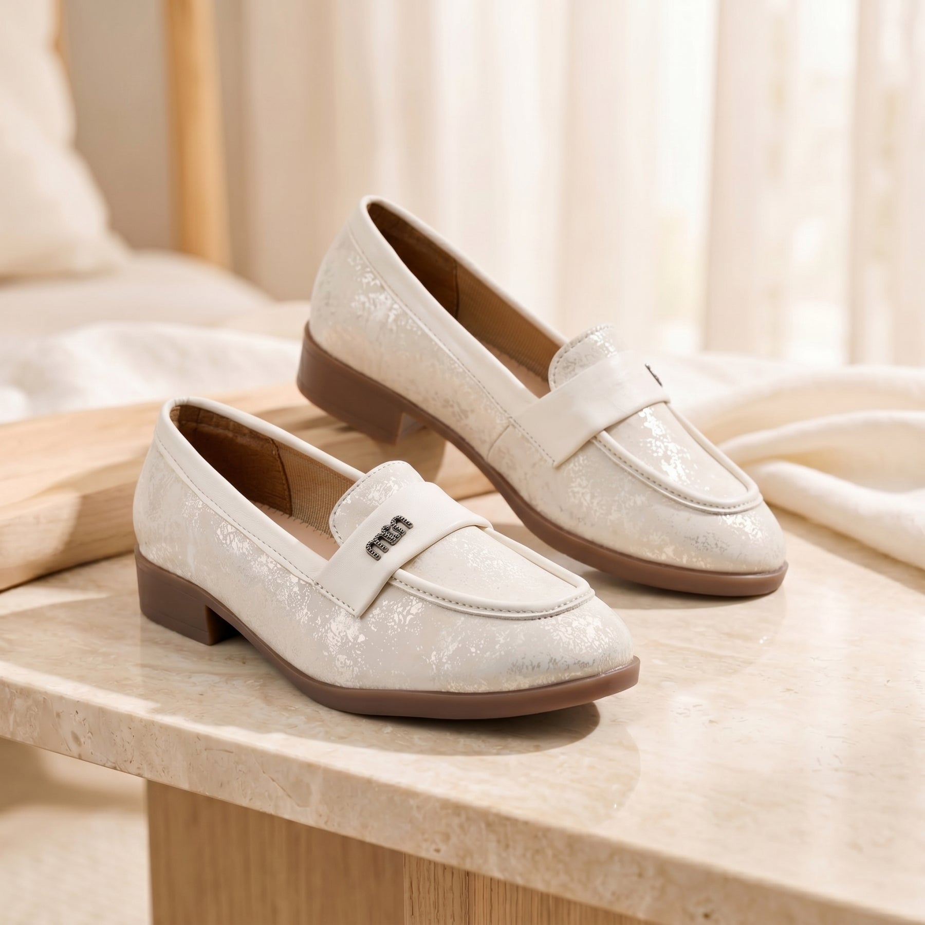 A pair of women's low-heeled loafers in a light cream or off-white color with a subtle textured pattern, resting on a light stone tabletop in a softly lit indoor setting.