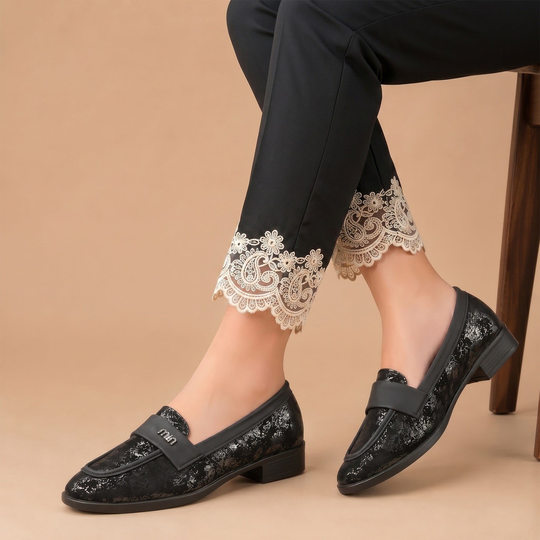 Black loafers with lace details worn with black pants on a beige background