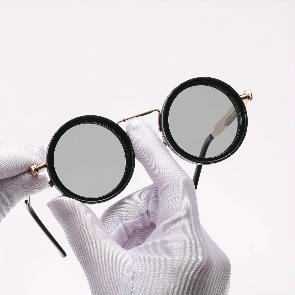 Round black-framed sunglasses with gray tinted lenses, held by a hand wearing a white glove against a plain white background.