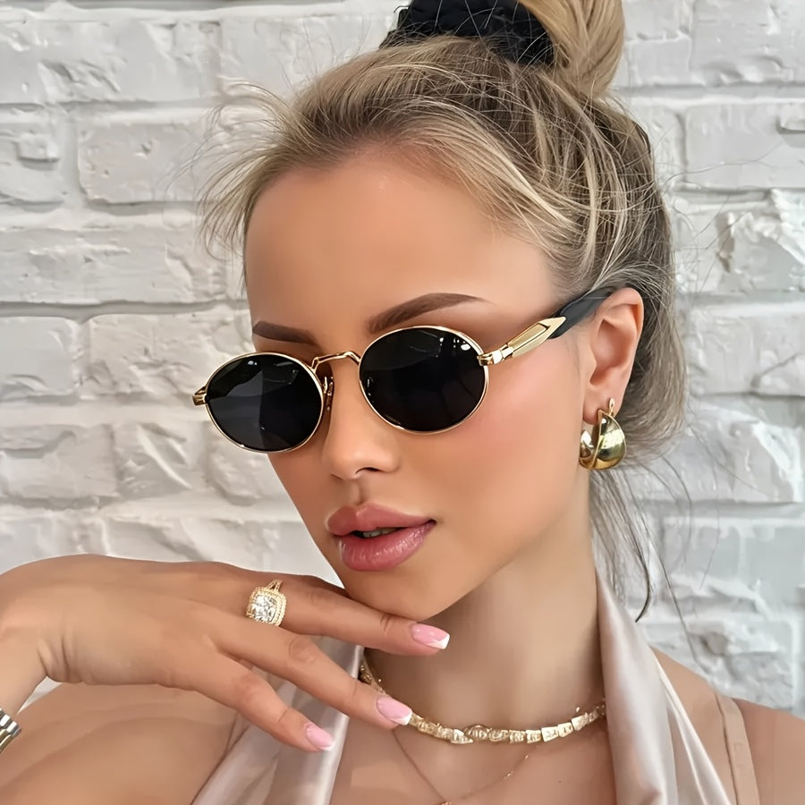 A close-up lifestyle shot of a woman wearing gold-framed oval sunglasses with dark lenses, styled with gold jewelry against a white brick wall background.