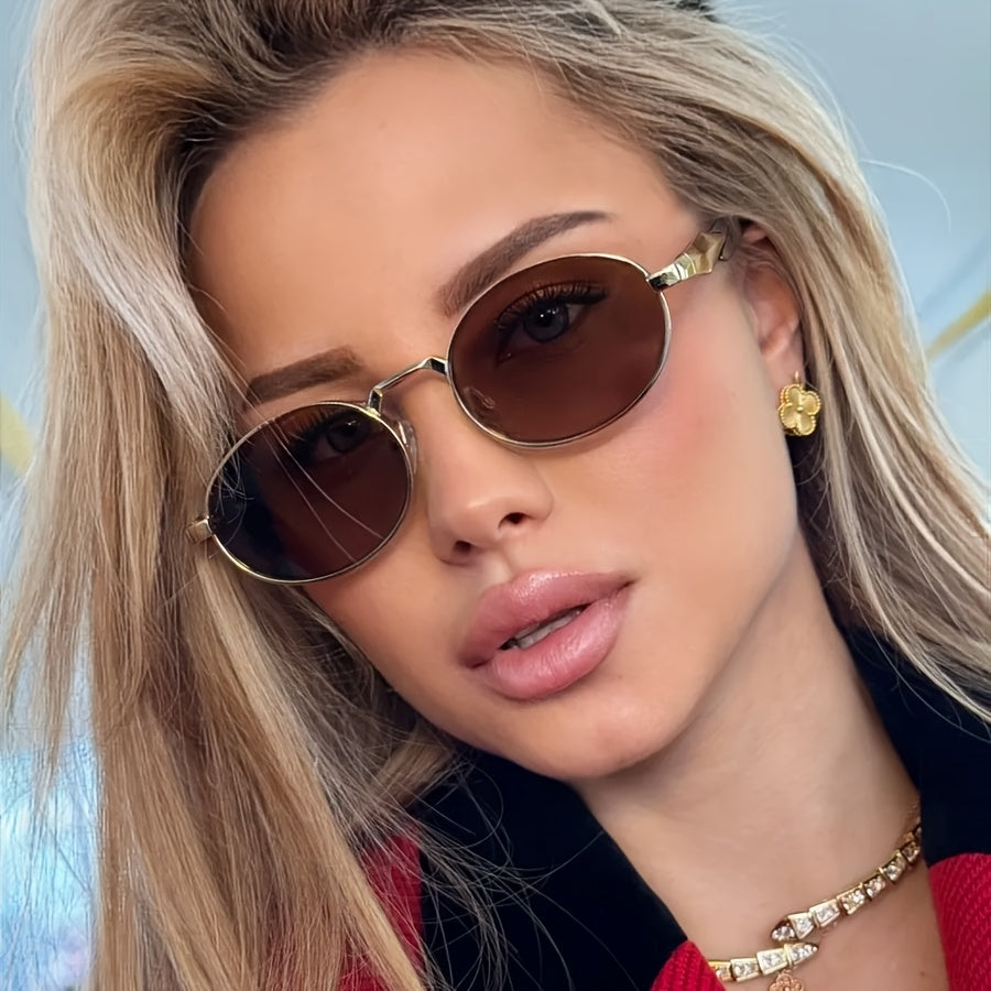 A close-up of a woman with blonde hair wearing stylish oval sunglasses with gold frames and dark brown lenses, along with a gold earring and necklace.