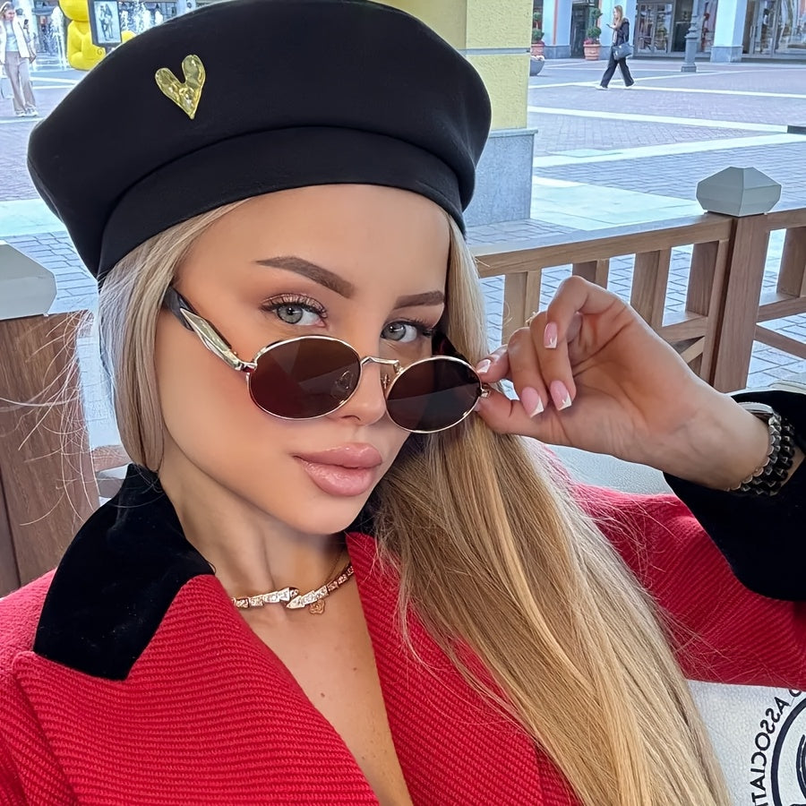 A close-up lifestyle shot of a woman wearing a black beret with a gold heart, gold-framed sunglasses, and a red jacket with a black collar, set against an outdoor plaza background.