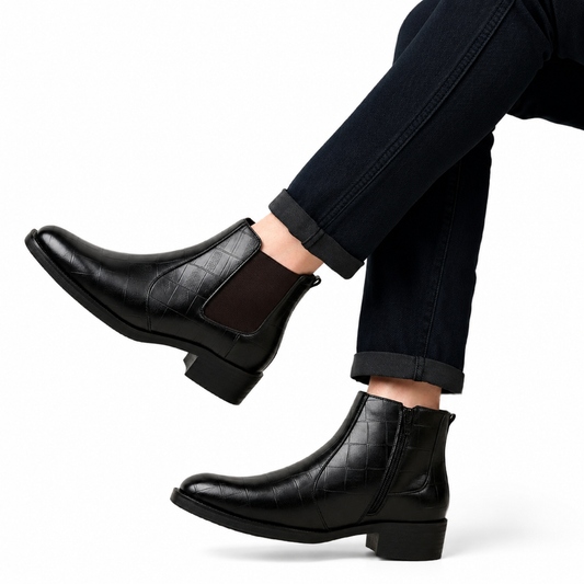 A pair of black leather ankle boots with a crocodile-embossed pattern and elastic side panels, shown on a model wearing dark cuffed jeans against a white background.