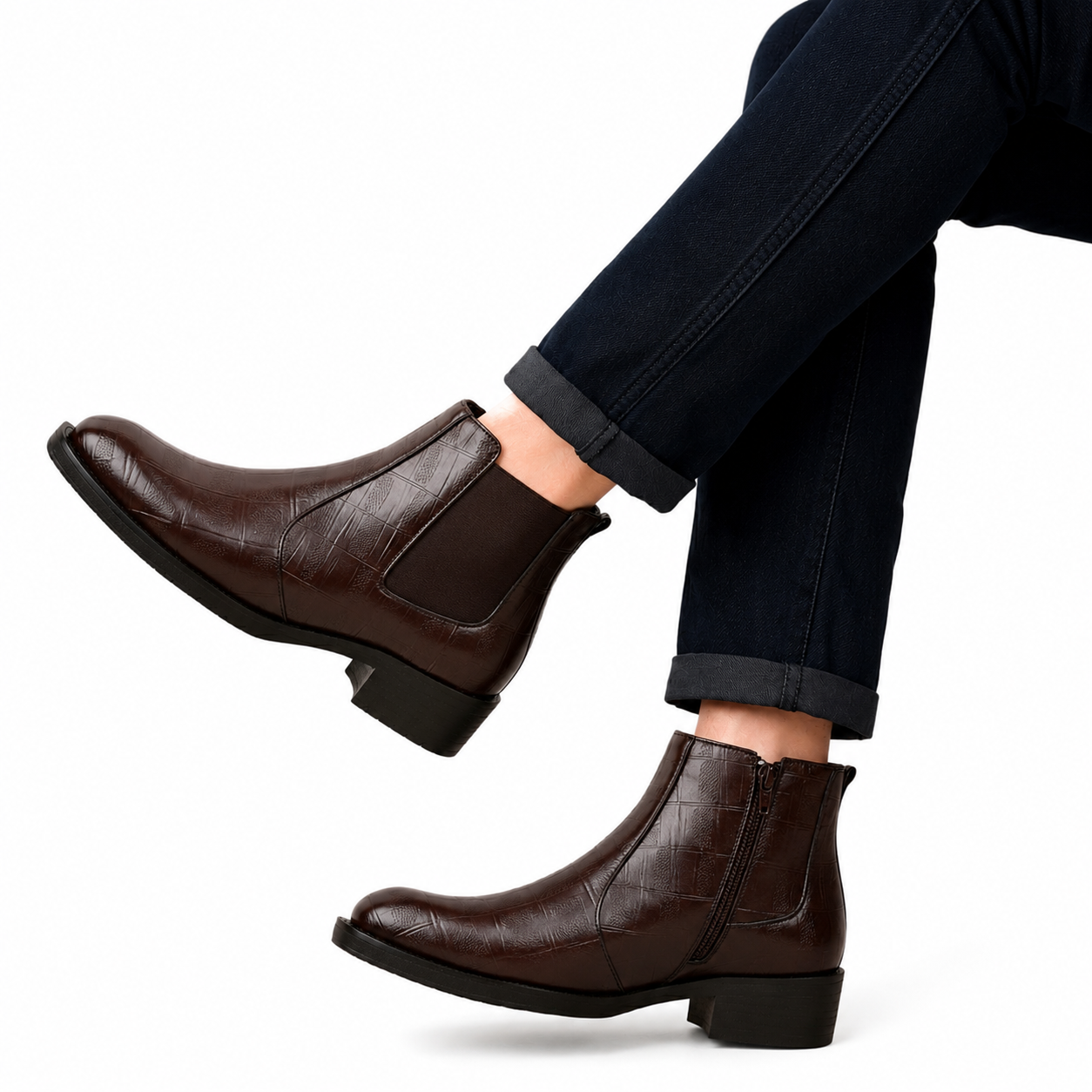 A pair of dark brown leather ankle boots with a crocodile-embossed texture and elastic side panels, shown on a model wearing dark cuffed jeans against a white background.