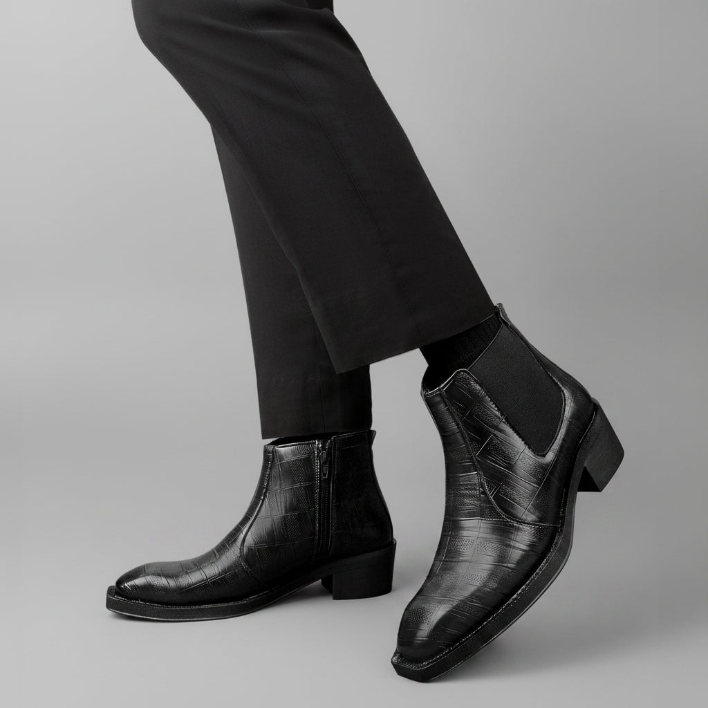 A pair of black leather ankle boots with a crocodile-embossed texture, shown on a model wearing black trousers against a solid brown background.