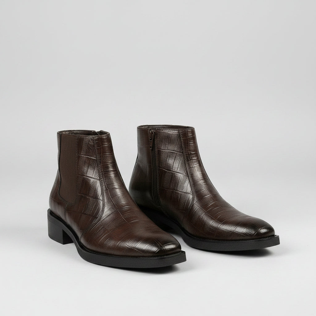 A pair of men's dark brown leather ankle boots with a crocodile-embossed texture, shown on a model wearing dark trousers against a warm beige background.