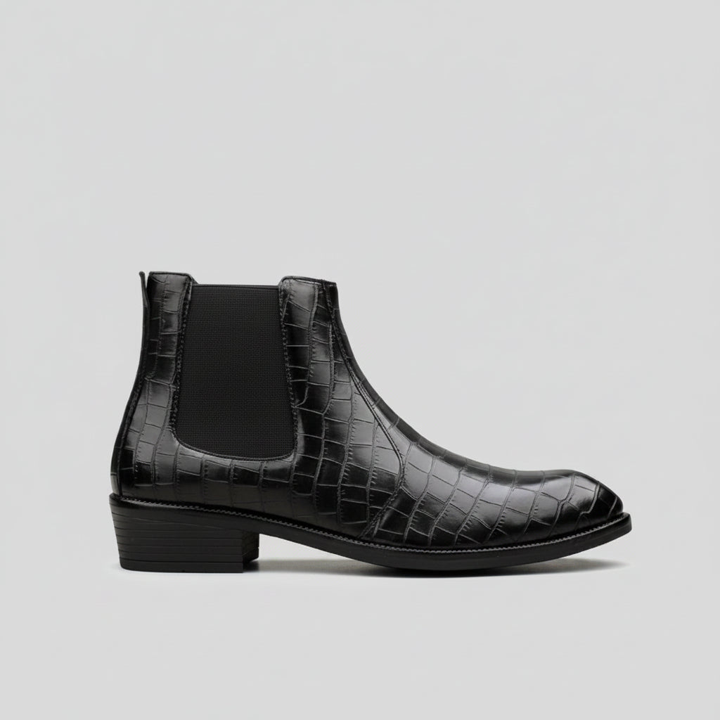 A single black ankle boot with a glossy crocodile-embossed texture and a black elastic side panel, shown in profile against a light gray background with blurred green plants.