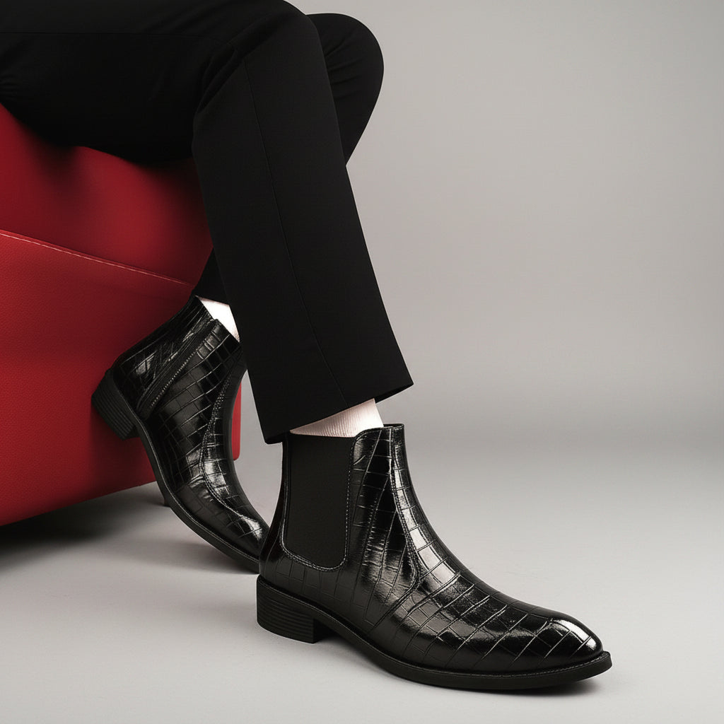 A person wearing black pointed-toe Chelsea ankle boots with a glossy crocodile-embossed texture and black elastic side panels, paired with black trousers and white socks, seated on a red chair against a brown wall and wood floor.