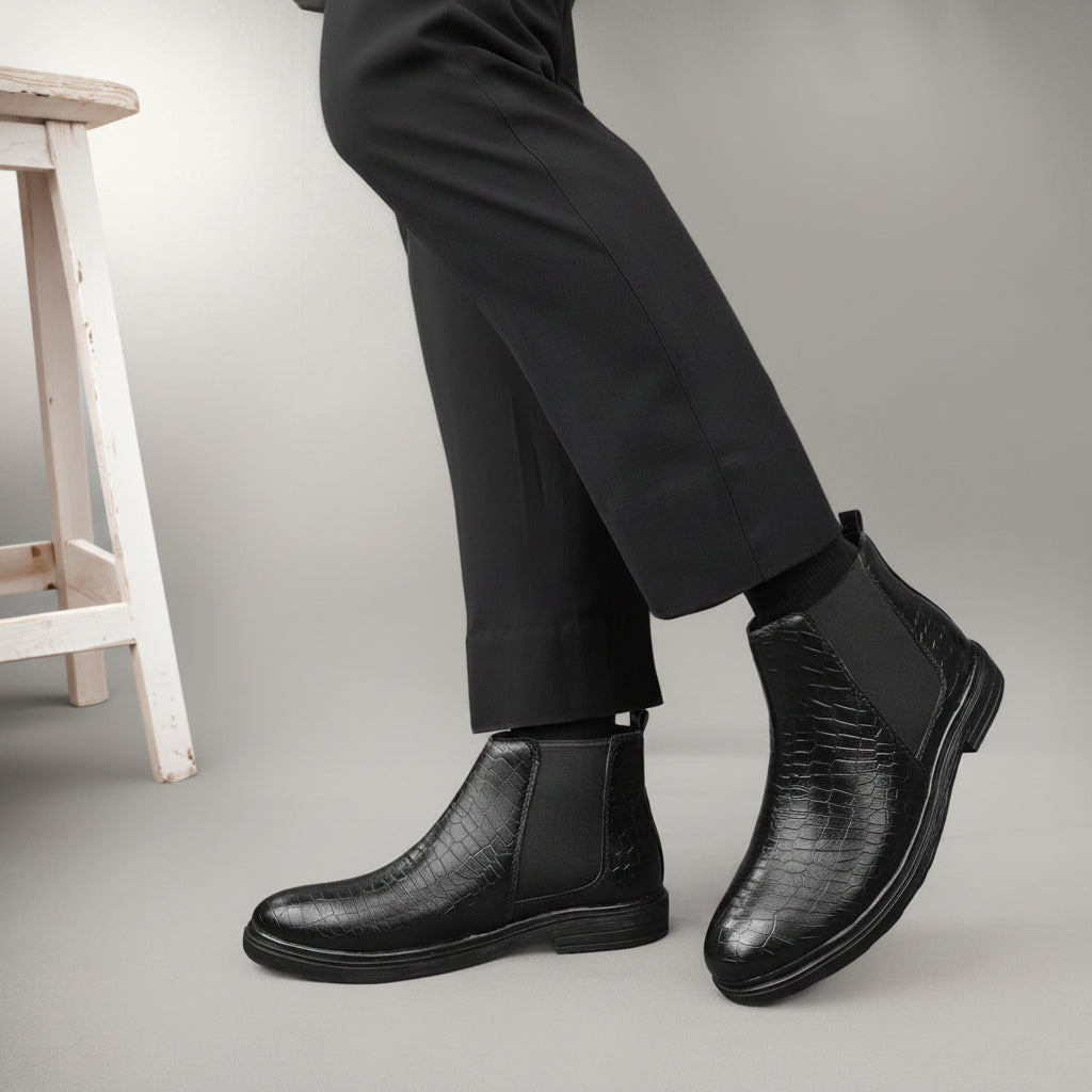 A person wearing black crocodile-embossed Chelsea boots with black elastic side panels, paired with black trousers, shown against a warm beige studio background with a white stool partially visible.
