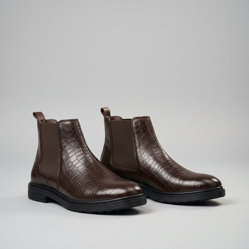 A pair of dark brown Chelsea boots with crocodile-embossed leather on the outer panels and elastic side panels, shown worn with navy trousers on a wooden floor.