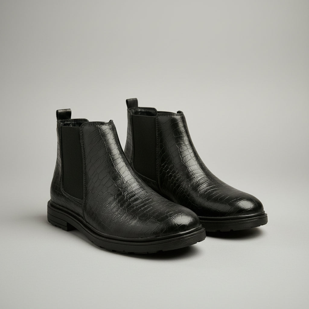 A pair of black ankle boots with a crocodile-embossed texture and elastic side panels, displayed on a wooden surface with rustic stone and leather background props.