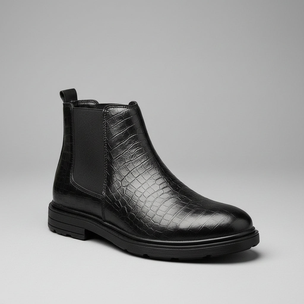 A single black ankle Chelsea boot with a crocodile-embossed texture on the toe and side, black elastic side panels, and a low stacked heel, resting on a wooden surface with a brown leather chair in the background.