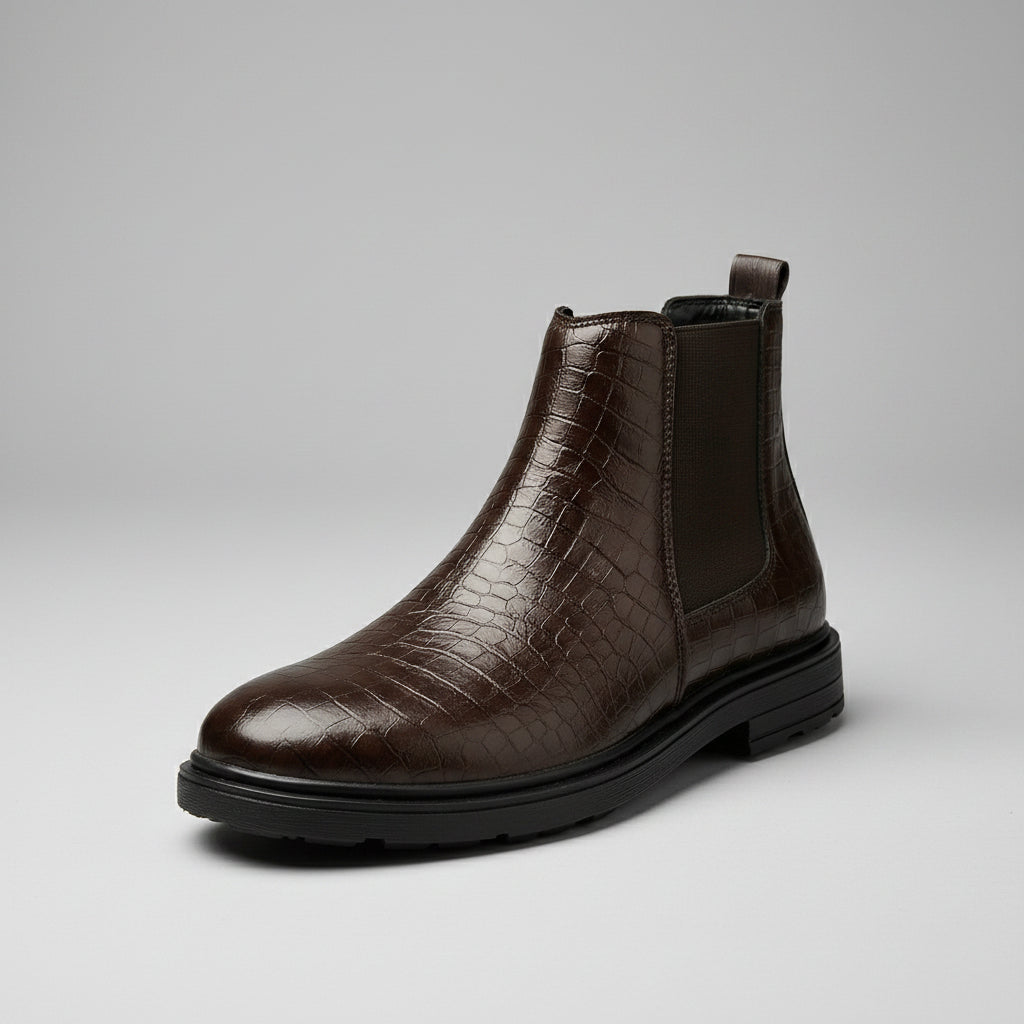 A dark brown Chelsea-style ankle boot with an embossed crocodile pattern on the front and side, a black elastic side panel, and a black sole, resting on a wooden table in a warm indoor setting.