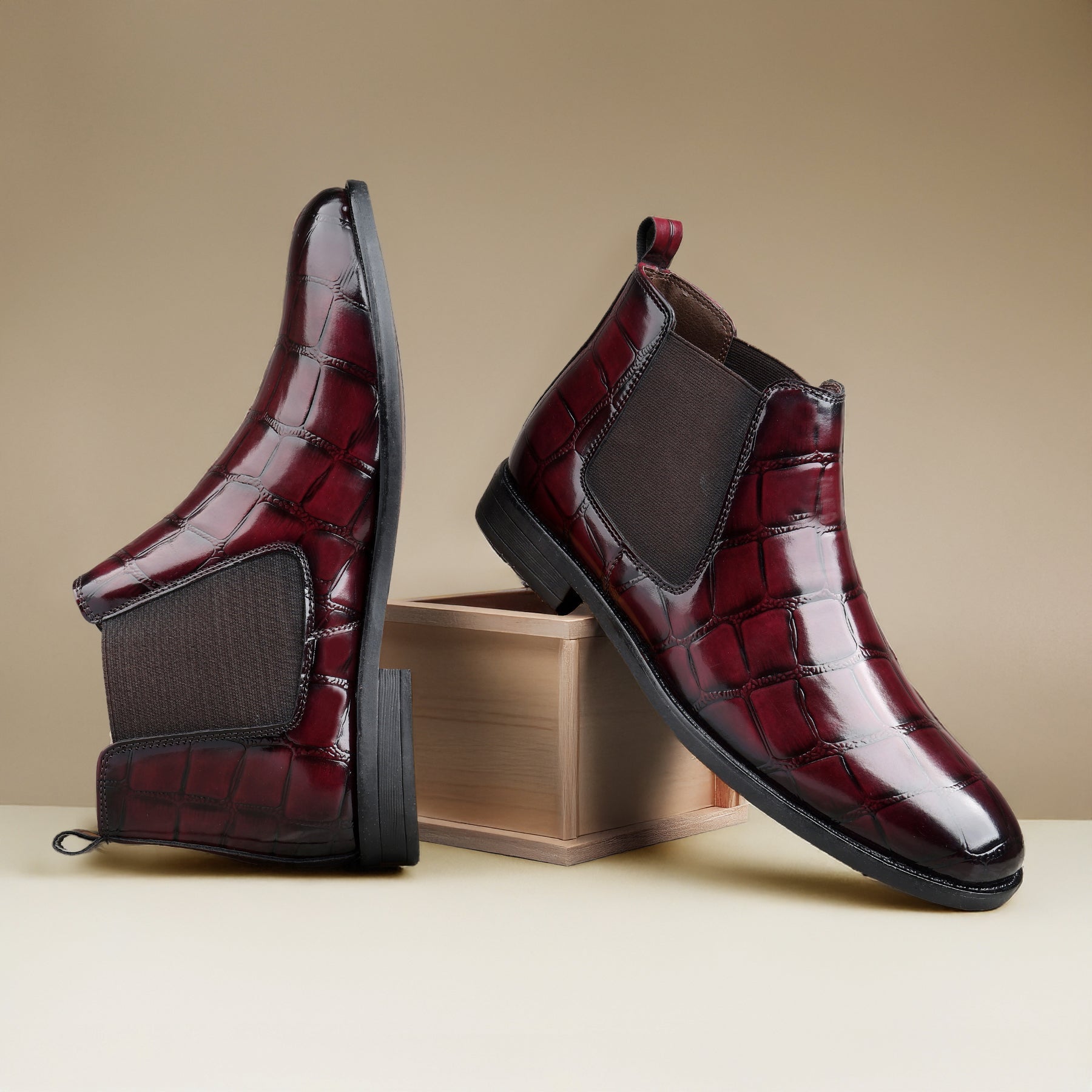 A pair of men's Chelsea-style ankle boots with a glossy crocodile-embossed leather upper in deep burgundy red, featuring dark brown elastic side panels and black soles. One boot is shown standing upright and the other is propped on a small wooden block against a warm beige studio background.