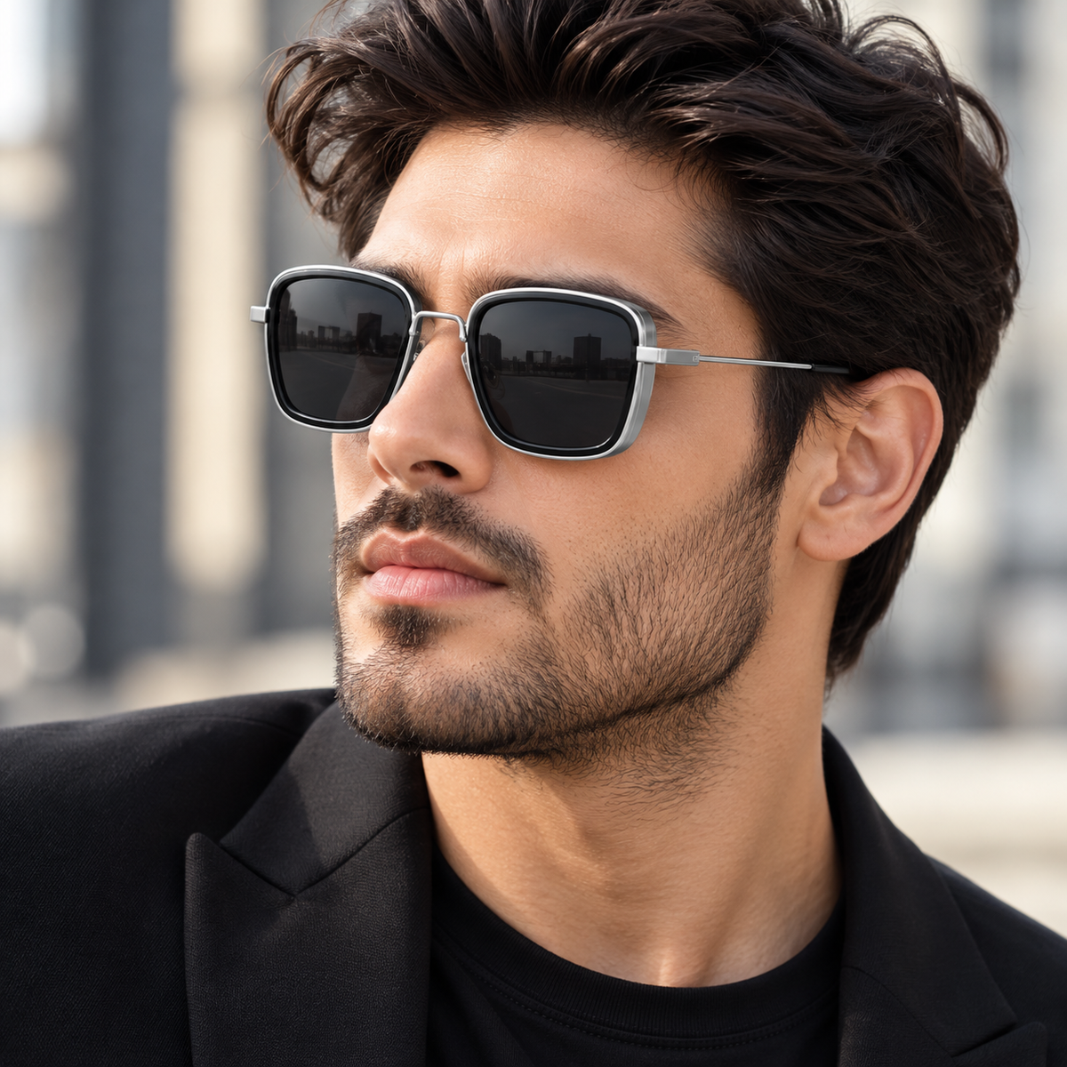 A close-up of a man wearing square sunglasses with a silver metal frame and dark lenses, dressed in a black blazer and t-shirt against a blurred outdoor background.