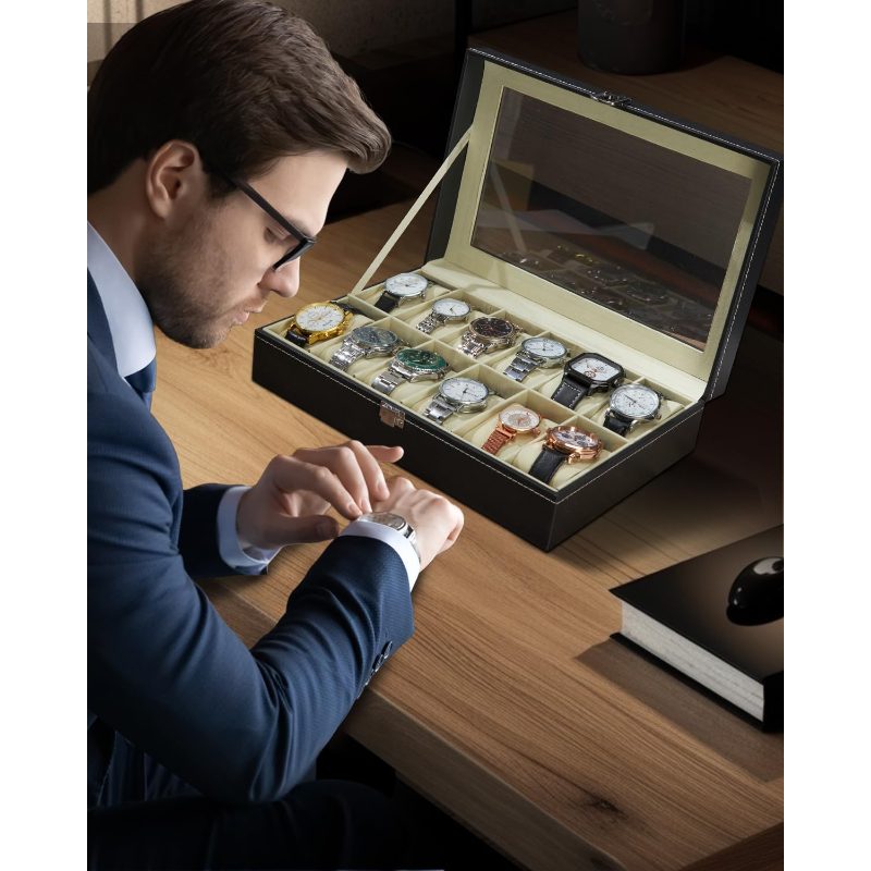A man in a navy blue suit and glasses watches examines a wristwatch while seated at a wooden desk next to an open luxury watch display case filled with assorted watches.
