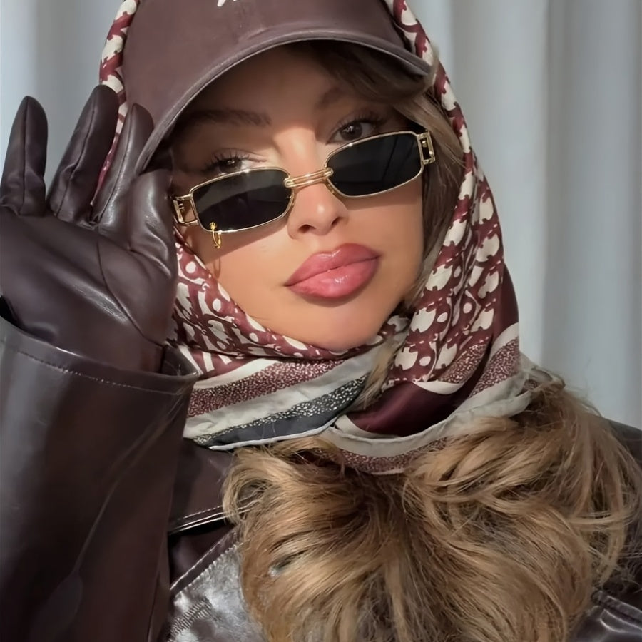 A close-up portrait of a woman wearing a brown leather cap, matching brown leather gloves, a brown and white patterned scarf, and gold-framed rectangular sunglasses.