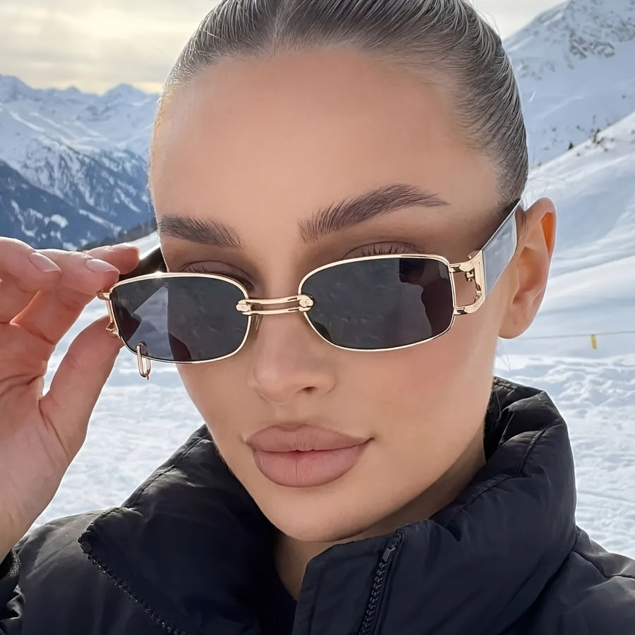 A close-up of a woman in a snowy mountain setting wearing gold-framed rimless sunglasses with dark lenses and a black puffer jacket.
