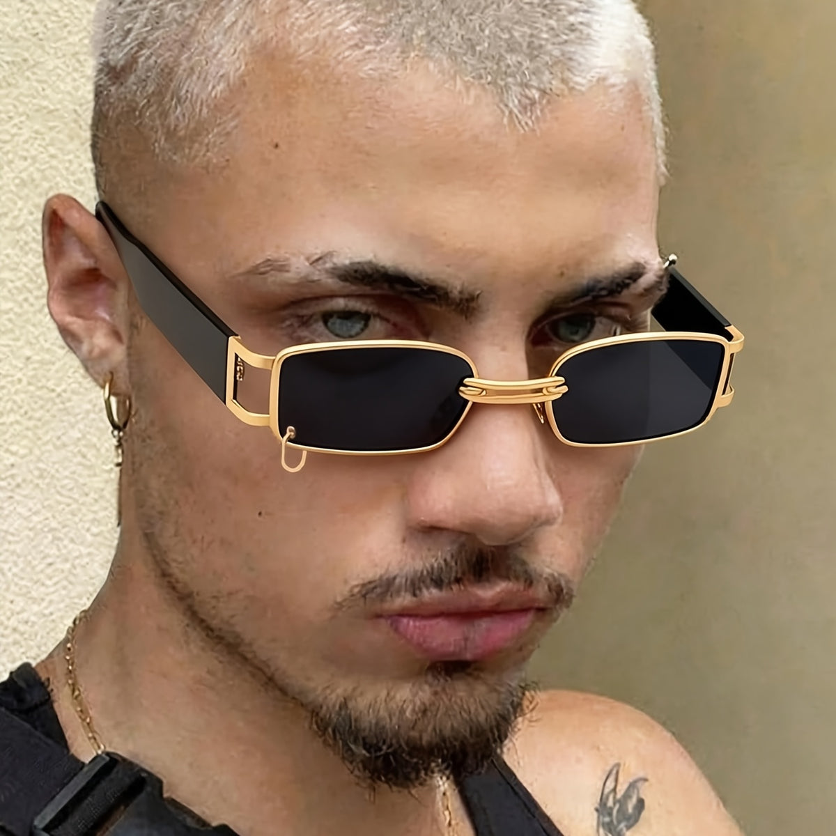 A close-up of a man wearing rectangular sunglasses with gold metal frames and black lenses. The sunglasses feature a unique small gold hanger detail on the left side of the frame.