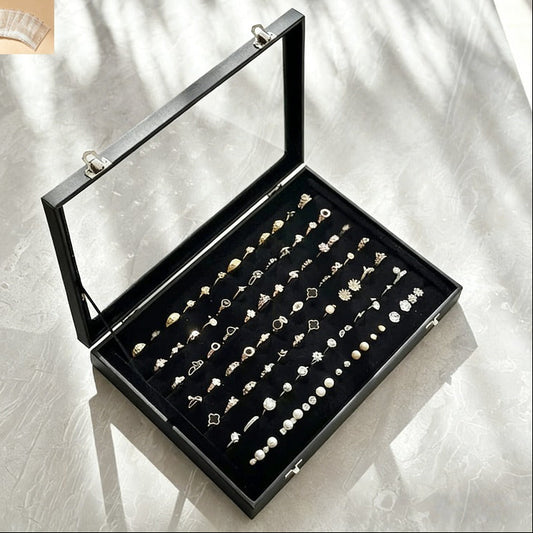 An open black jewelry box with a clear glass lid, displaying dozens of rings on a black insert. The box is photographed on a light marble-like surface with soft window shadows.