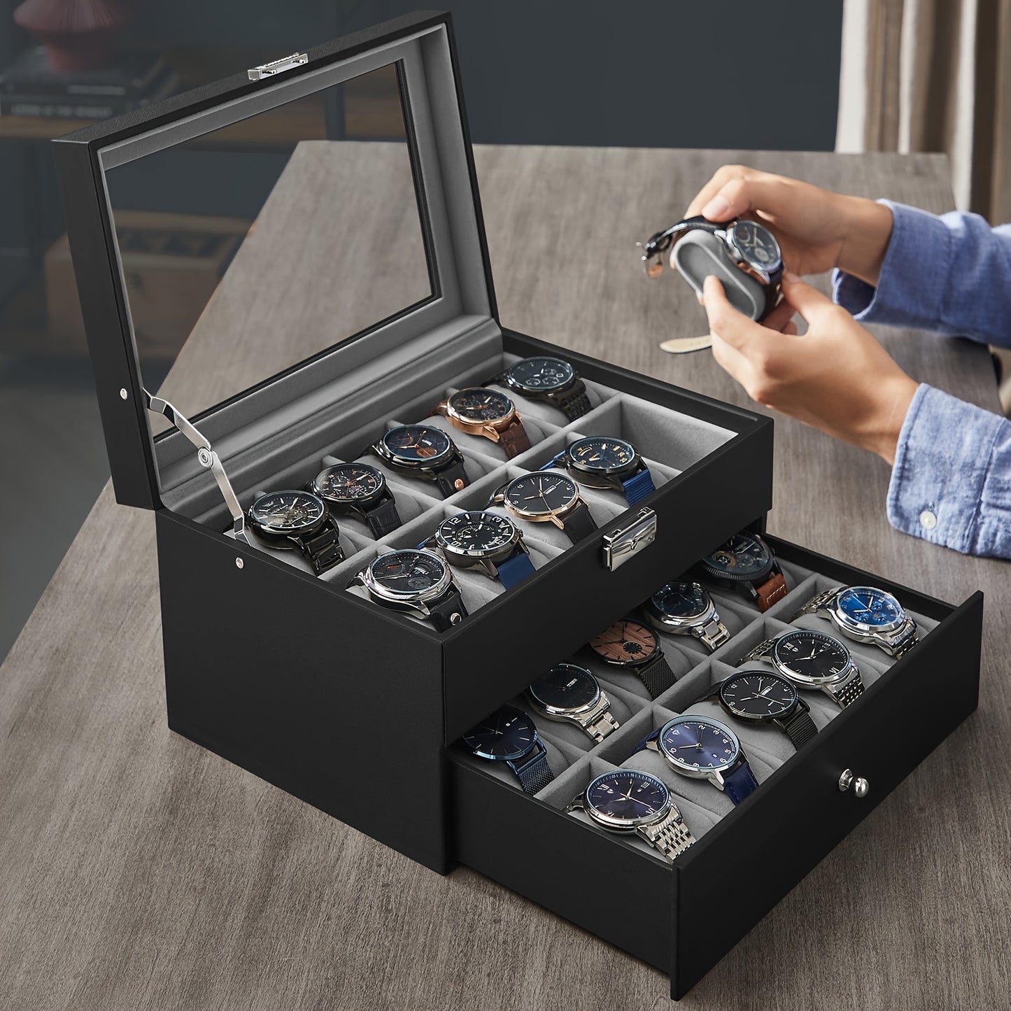 A black watch box with a glass lid and two drawers, shown open on a wooden table. Several wristwatches are stored inside, and a person's hands are visible holding one of the watches above the box.