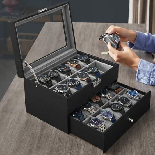 A black watch box with a glass lid and two drawers, shown open on a wooden table. Several wristwatches are stored inside, and a person's hands are visible holding one of the watches above the box.