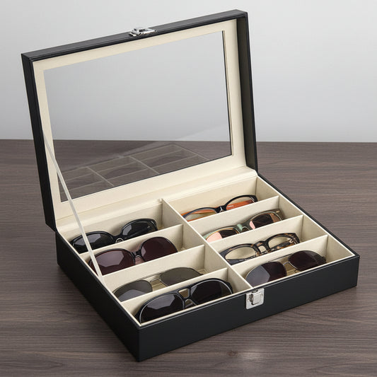 A black faux-leather sunglasses storage box with a clear glass lid and an interior mirror, shown open with six compartments holding various sunglasses. The box has a beige interior lining and silver metal latches.