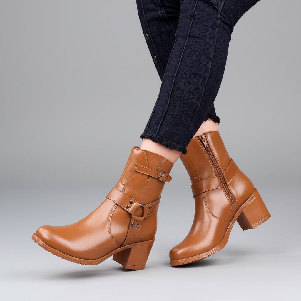 A person wearing tan ankle boots with side zippers and buckle straps, paired with dark jeans, seated on a wooden stool against a solid green background.