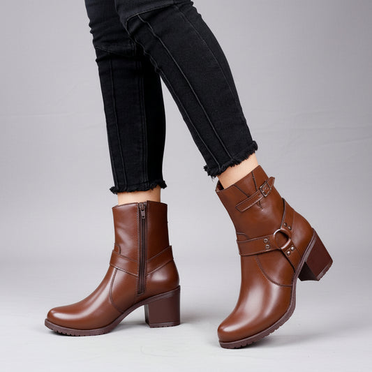 A person wearing brown ankle boots with side zippers and buckle strap details, paired with black jeans, standing against a gray concrete wall.