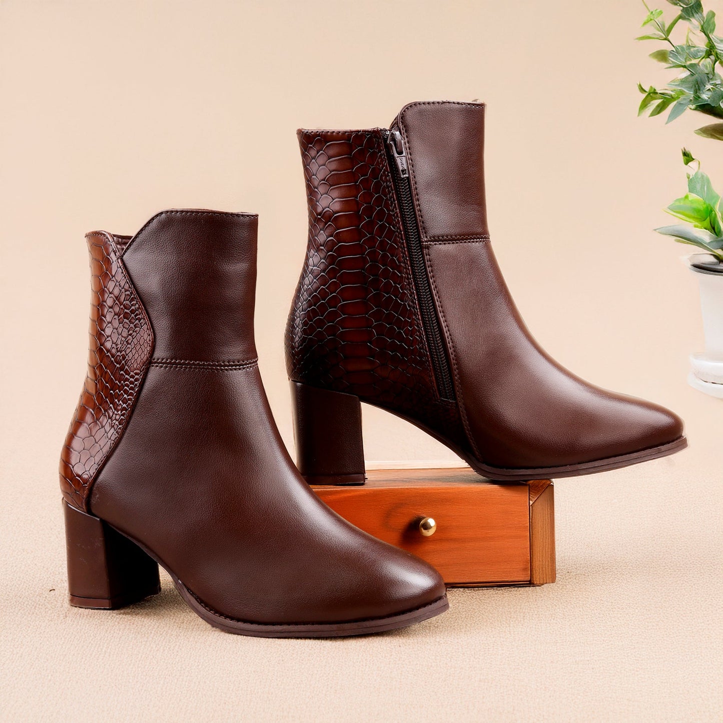 A pair of dark brown ankle boots with a mix of smooth leather and crocodile-embossed textures, displayed on a small wooden block against a beige background.