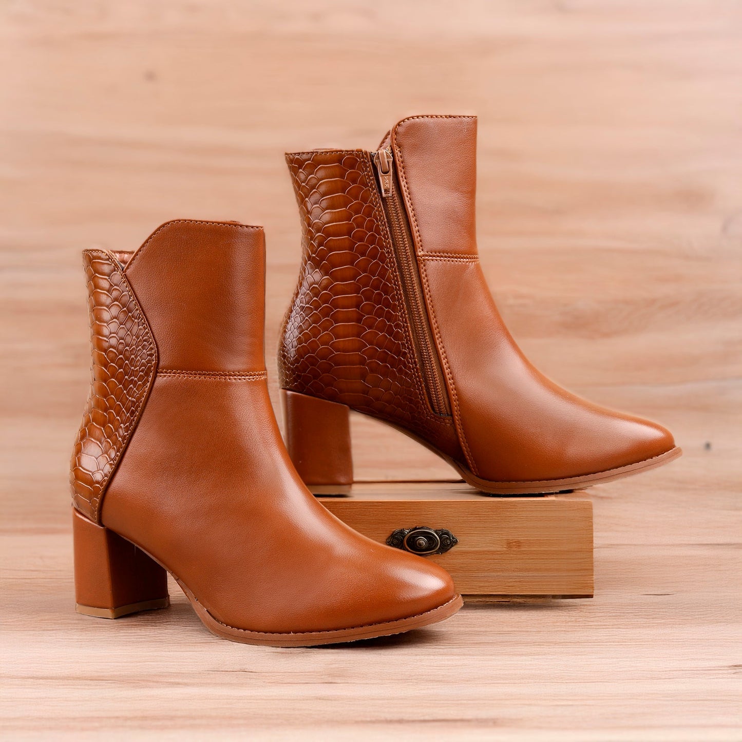A pair of tan leather ankle boots with a block heel, shown on a light wood background. One boot is positioned in front on a small wooden block, and the other stands behind it.
