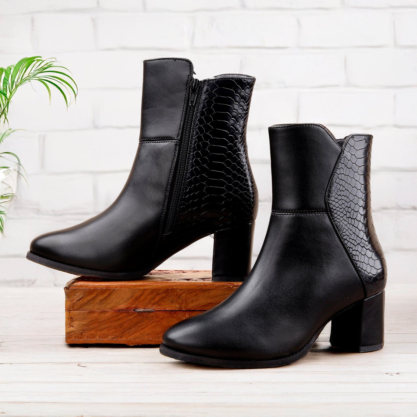 A pair of black ankle boots with a mix of smooth and crocodile-embossed textures, displayed on a wooden block against a white brick wall background.