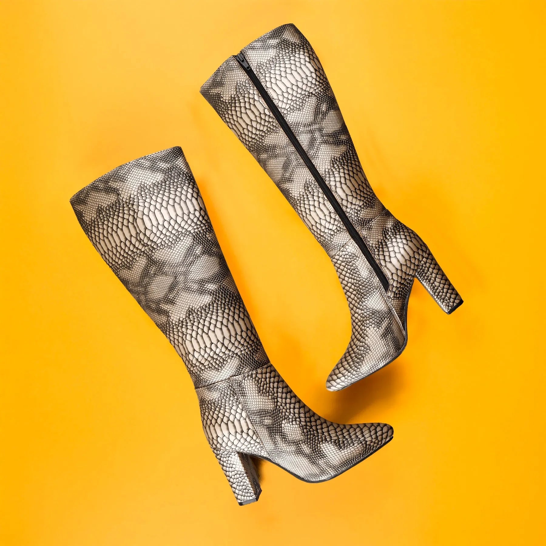 A pair of knee-high pointed-toe boots with a black and gray snakeskin pattern, shown from an overhead flat-lay perspective on a solid bright yellow background.