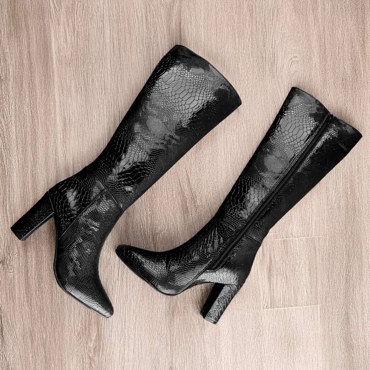 A pair of black knee-high boots with a glossy crocodile-embossed texture, featuring pointed toes and block heels, displayed on a light wood floor.