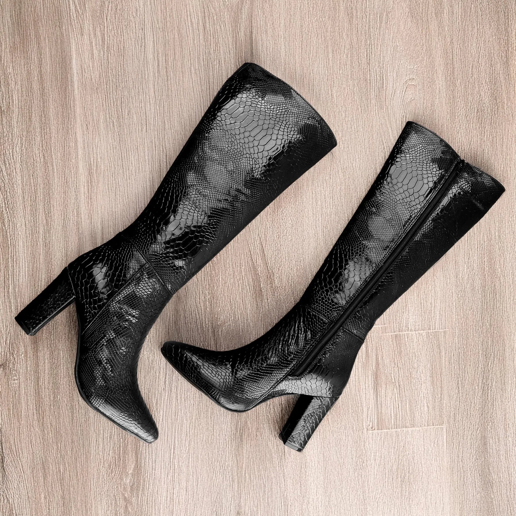 A pair of black knee-high boots with a glossy crocodile-embossed texture, featuring pointed toes and block heels, displayed on a light wood floor.