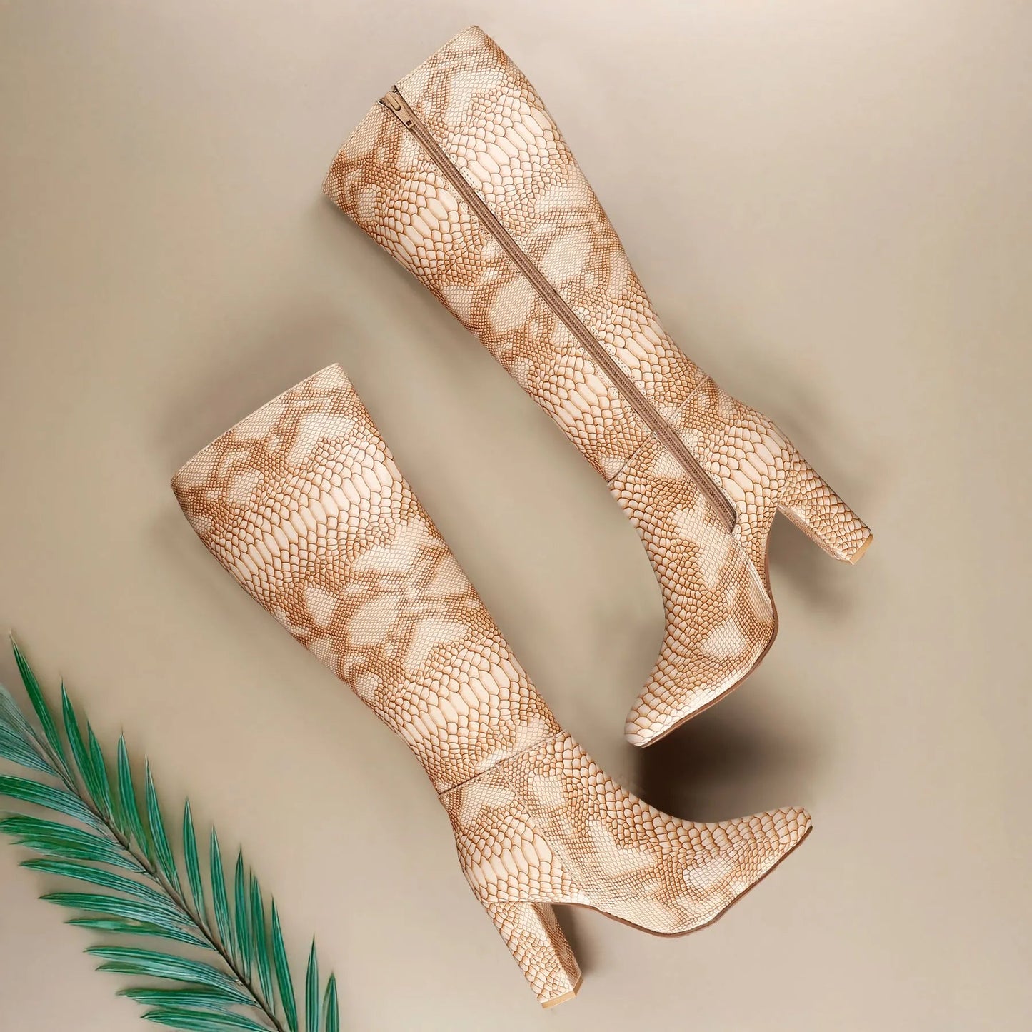 A pair of knee-high pointed-toe boots with a beige and white snakeskin pattern, featuring a high block heel and side zipper, displayed on a neutral beige background with a green palm leaf prop.