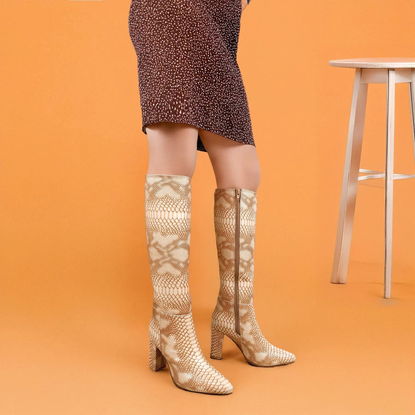 A model's legs wearing knee-high snakeskin print heeled boots, paired with a brown patterned mini skirt, set against a solid orange background with a wooden stool on the right.