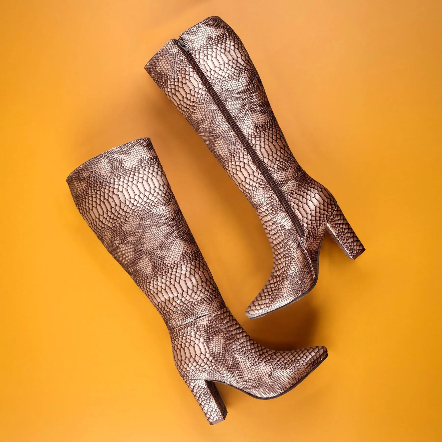 A pair of knee-high heeled boots with a metallic snakeskin pattern in bronze and brown, arranged diagonally on a solid orange background.