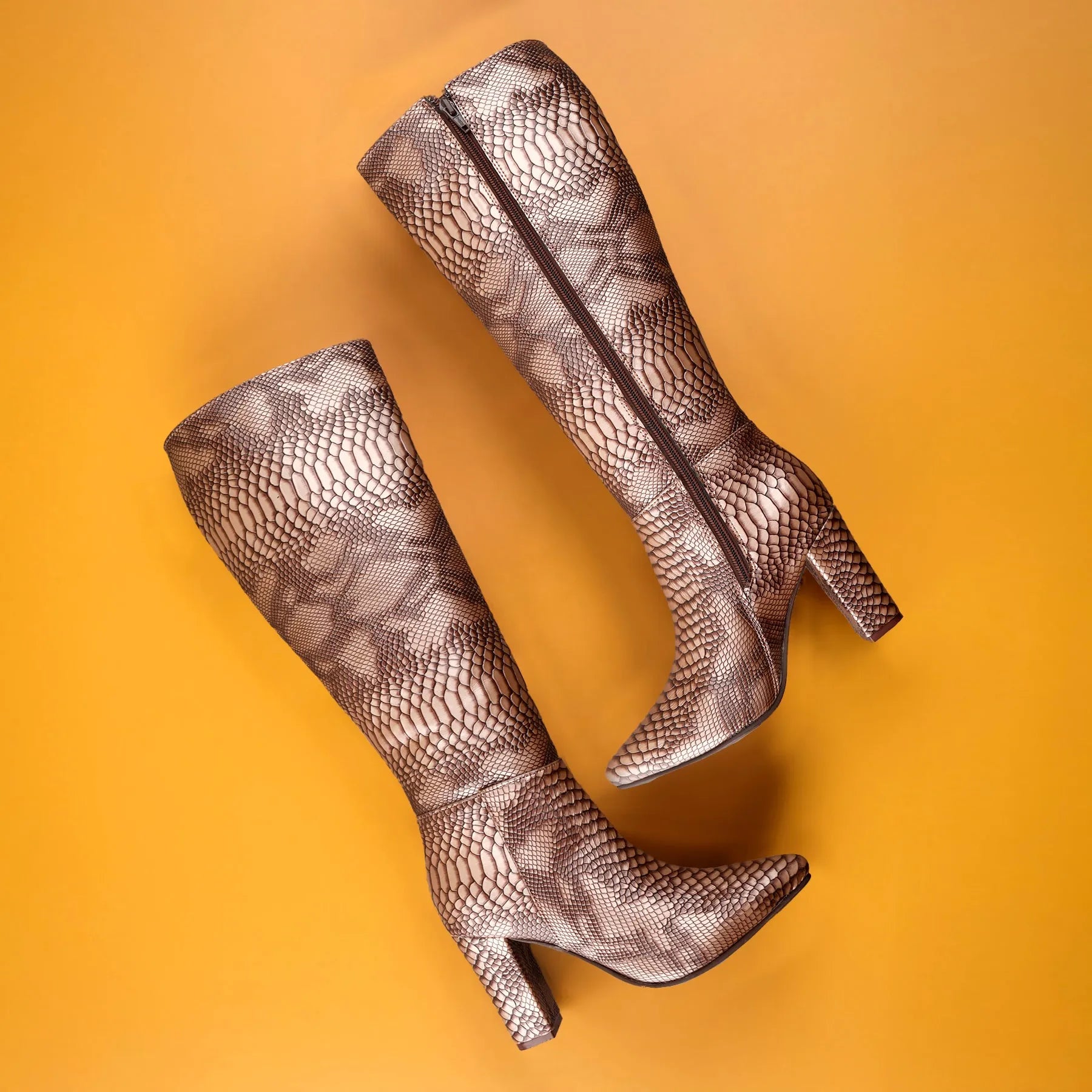 A pair of knee-high heeled boots with a metallic snakeskin pattern in bronze and brown, arranged diagonally on a solid orange background.