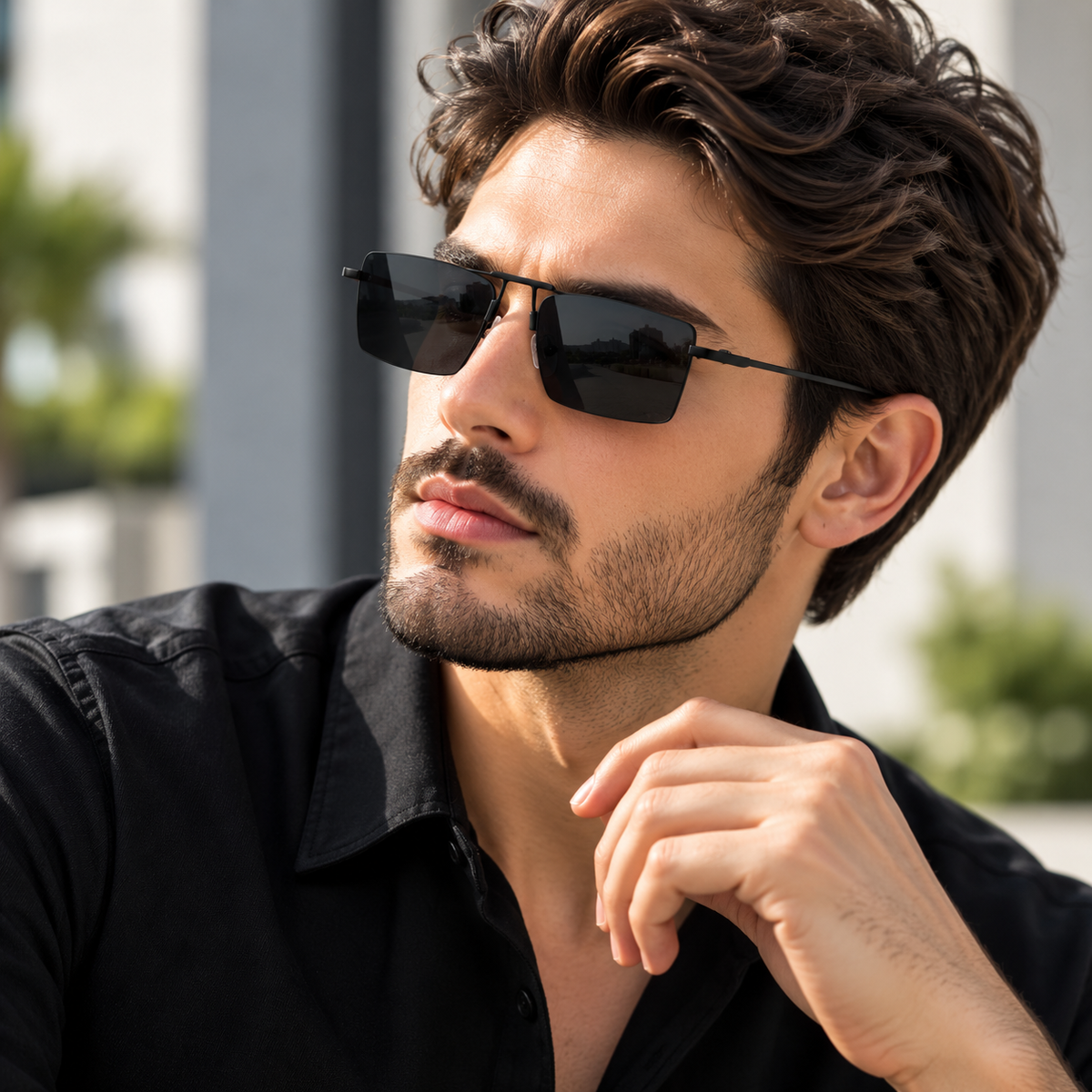 A man with styled brown hair and a beard wearing black square sunglasses and a black button-up shirt, photographed outdoors with a blurred light gray building background.