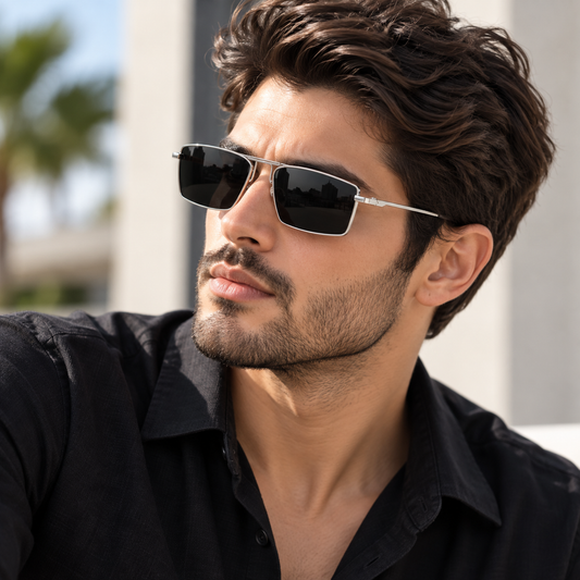 A man wearing square aviator-style sunglasses with dark lenses and thin silver metal frames, dressed in a black collared shirt against a softly blurred outdoor background.