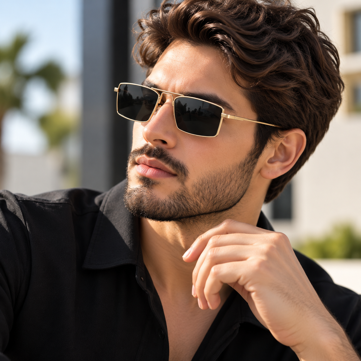 A man wearing gold-framed square sunglasses with dark lenses, posing outdoors in a black collared shirt.
