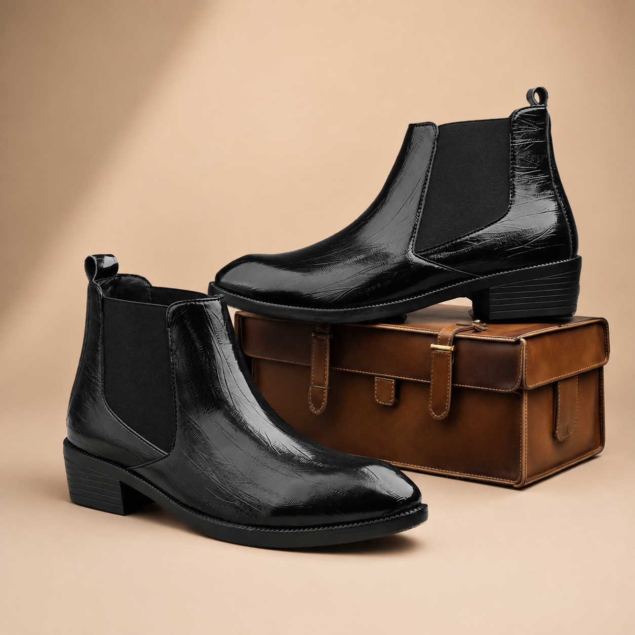 A pair of black Chelsea-style ankle boots with a glossy finish and low block heels, displayed on and around a brown leather suitcase against a warm beige studio background.