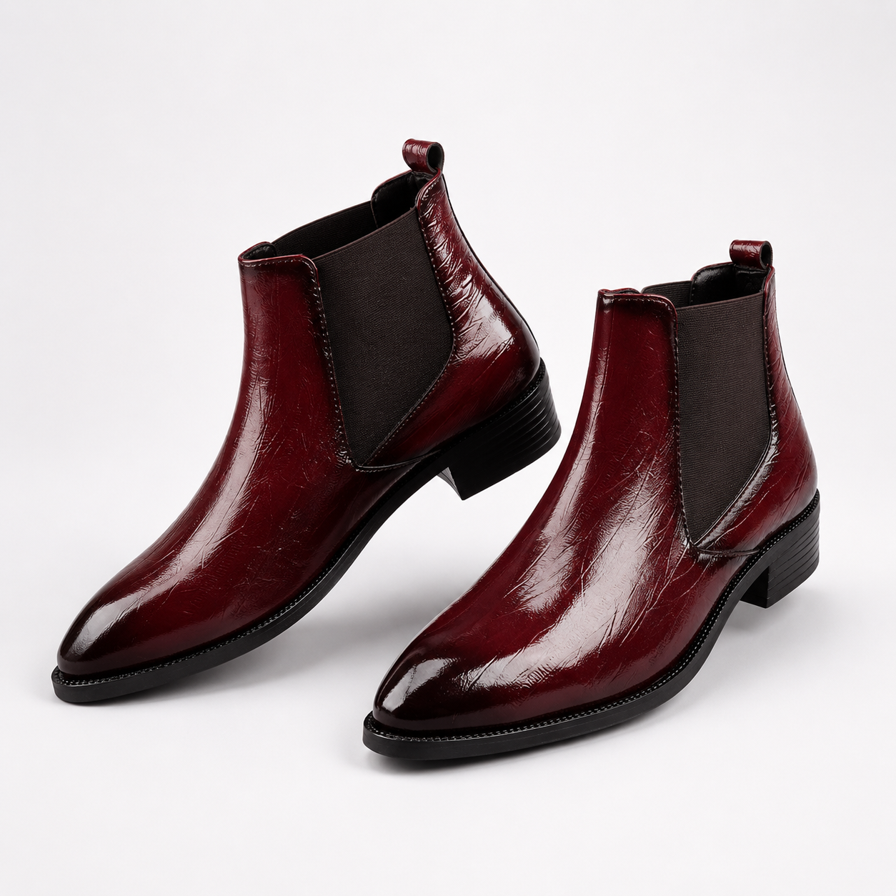 A pair of glossy burgundy leather Chelsea ankle boots with black elastic side panels and low black block heels, photographed on a clean white studio background.